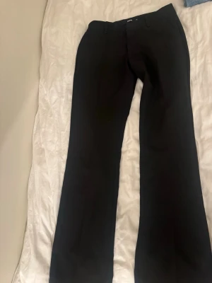 Svarta kostymbyxor från Bikbok - Svarta kostymbyxor från Bikbok i klassisk rak modell med bälteshällor och diskreta fickor både fram och bak. Byxorna har en lowwaist-passform och är gjorda i ett mjukt material som känns skönt mot huden. Perfekt för en clean och stilren look.