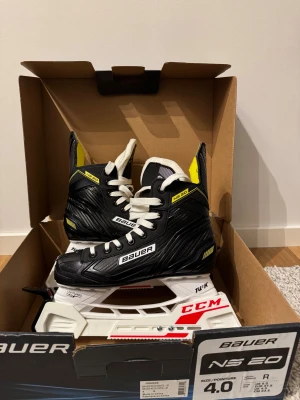 Svarta Bauer NS 20 skridskor - Grymma Bauer NS 20 skridskor i svart med vita snören och detaljer i gult. Yttermaterialet har en kolfiberlook och innersidan är vadderad för extra komfort. Skenorna är i rostfritt stål och hållarna är vita med röda CCM-detaljer. Storlek 37,5 - 38. Skridskorna är använda ca 2 gånger och är nyskick! Hör av dig vid minsta intresse!😊
