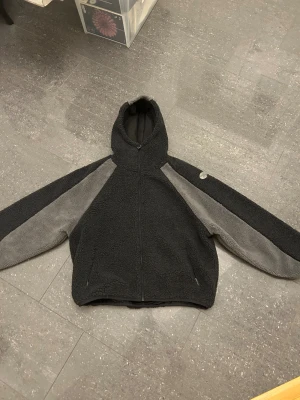 Mertra yeti fleece - Yeti fleece med huva från Merta i storlek XL. Jackan är svart med gråa paneler längs ärmar och axlar, hel dragkedja framtill och två fickor. Mysigt teddy-material som håller dig varm och snygg under kyliga dagar.