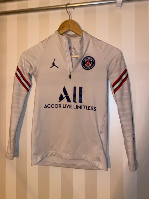 Vit långärmad PSG träningstopp Jordan - Säljer en vit långärmad träningstopp från Jordan och PSG med halv dragkedja framtill. Toppen har blå och röda ränder på ärmarna, Paris Saint-Germain-logga på bröstet och tryck på ärmen. Materialet är mjukt och syntetiskt, perfekt för sport.