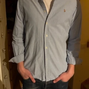 Ljusblå skjorta från Ralph Lauren - Säljer en ljusblå skjorta från Ralph Lauren i klassisk passform. Skjortan har knappar framtill, lång ärm och den ikoniska loggan broderad på bröstet. Perfekt för dig som gillar stilrena och tidlösa plagg. Materialet är mjuk bomull.