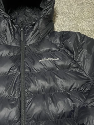 Svart Peak Performance dunjacka - Säljer en svart dunjacka från Peak Performance med huva och ribbade muddar. Jackan har en snygg quiltad design, dragkedja framtill och logga broderad på bröstet och ärmen. Perfekt för kalla dagar och har en modern, loose passform.