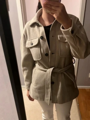 Zara kappa  - Säljer en beige overshirt från Zara i storlek M eftersom den inte passar mig bra. Jackan har två stora bröstfickor med knappar, klassisk krage och knäppning framtill. Den har ett matchande bälte i midjan. Skriv privat för fler frågor/bilder