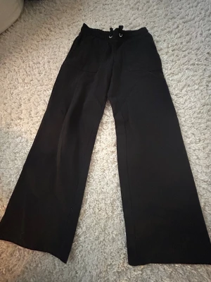 Svarta wide byxor från 157 - Säljer ett par svarta wide byxor från 157 med snörning i midjan och stora fickor framtill. Byxorna har en avslappnad passform och är perfekta för dig som gillar en loose stil. Materialet känns mjukt och bekvämt mot huden.