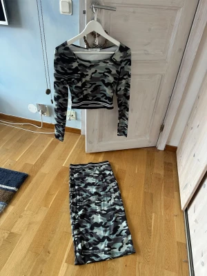 Camouflage crop top set från Voyelles - Säljer en långärmad crop top och kjol från Voyelles i coolt camouflage-mönster. Toppen har rund halsringning och ribbade svarta och vita ränder i nederkant. Materialet är stretchigt och mjukt, perfekt för en edgy streetstyle-look.