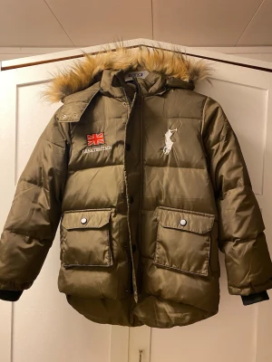 Olivgrön pufferjacka med pälsluva - Olivgrön pufferjacka från ProJob med stor huva och fluffig fuskpäls. Jackan har broderade detaljer med brittisk flagga och texten 'Great Britain' samt en vit polospelare på bröstet. Två stora fickor framtill med knappar och dragkedja. Perfekt för kalla dagar. Ärm längd 50 cm bröst mått 46 cm längd 55 cm fram 65 cm baksida.