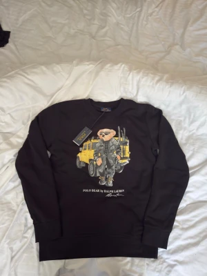 Polo Bear Ralph Lauren svart sweatshirt  - Svart sweatshirt från Polo Ralph Lauren med coolt Polo Bear-motiv framtill där björnen står framför en gul bil och har solglasögon och jacka. Rund halsringning och långa ärmar. Mjuk bomullskvalitet och casual vibe, perfekt för chill dagar. PRIS KAN DISKUETARS
