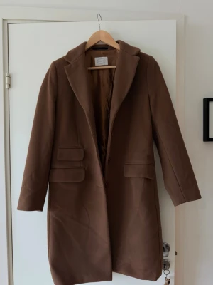 Elegant brun ullkappa från Newhouse - Kappa från Newhouse i en varm brun/kamel nyans i storlek XS. Klassisk rak modell med slagkrage, 4 knappar och fickor framtill. Kappan är i fint skick.
