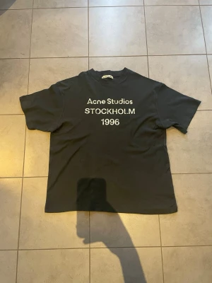 Svart Acne Studios t-shirt Stockholm - Säljer en svart t-shirt från Acne Studios med tryck 'Acne Studios STOCKHOLM 1996' på bröstet. Klassisk rund hals och korta ärmar. Skön passform och tillverkad i mjuk bomull. Perfekt för dig som gillar minimalistisk och stilren design.