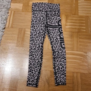 Leopardmönstrade leggings Aim'n XS - Snygga leopardmönstrade leggings från Aim'n i storlek XS. De är högmidjade och har ett svart tryck längs benet. Materialet är stretchigt med 73% polyester och 27% spandex, vilket gör dem perfekta för träning eller chill. Färgen är ljusgrå med svarta leopardfläckar.