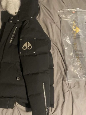 Svart pufferjacka från Moose Knuckles - Säljer en svart pufferjacka från Moose Knuckles med fet vit päls på huvan och coola metalldetaljer. Jackan har dragkedjor på ärmar och bröst, samt knappar framtill. Loggan sitter på ärmen och jackan har en schysst oversized look.