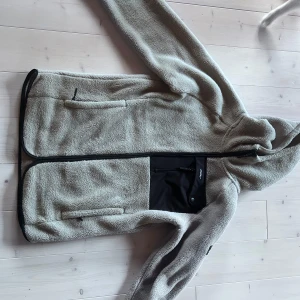 Beige teddyjacka från Revolution - Mysig beige teddyjacka med huva från Revolution. Jackan har svart bröstficka med dragkedja, två sidofickor och svarta detaljer längs kanterna. Perfekt för kyliga dagar och har en avslappnad passform med långa ärmar. Använd 1 gång