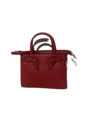 Röd handväska med rosettdetaljer - Snygg röd handväska i skinn med två handtag och avtagbar axelrem. Framsidan har två dekorativa rosettdetaljer som ger väskan en unik look. Perfekt storlek för det viktigaste och enkel att matcha med olika outfits.