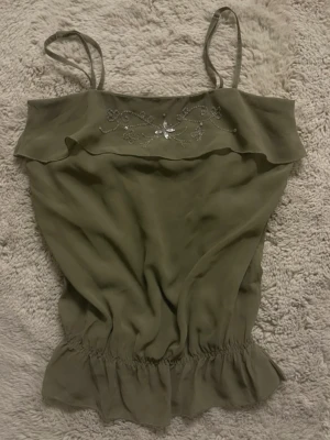 Olivgrön linnetopp med pärlor - Säljer en olivgrön linnetopp med smala axelband och dekorativa pärlor och paljetter i blommönster upptill. Toppen har volangkant vid bysten och ett smockat parti längst ner som ger en snygg form. Materialet är lätt och luftigt, perfekt för varma dagar.