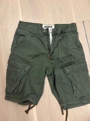 Gröna cargo shorts - Säljer ett par vintage gröna cargo shorts från Dcate i storlek XS. De har flera stora fickor med lock, tryckdetaljer på ena benet och dragkedja fram. Pris går att diskutera💓💓