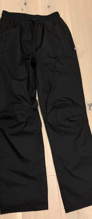 Svarta täckbyxor  - Helt nya svarta täckbyxor från Trekmates i storlek XS. Byxorna har elastisk midja med snörning, raka ben och är gjorda i ett slitstarkt syntetmaterial som står emot väta. Perfekta för kalla och blöta dagar utomhus. 