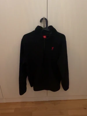 Svart fleece-tröja med half zip - Svart fleece-tröja från Liverpool FC med halv dragkedja och rött klubbmärke broderat på bröstet. Tröjan har lång ärm och en hög krage som ger extra värme. Perfekt för kyliga dagar och för dig som gillar fotboll.