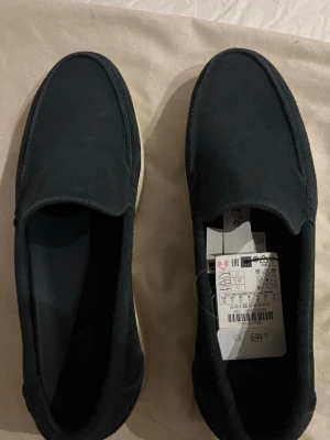 Svarta mockaloafers med vit sula - Snygga svarta loafers från H&M i mjuk mocka med vit platt sula. Skorna har en stilren design utan snörning och passar perfekt till jeans eller chinos. Enkla att dra på och av, med diskreta sömmar och klassisk rund tå.