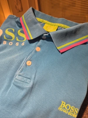 Torkus Blå pikétröja Hugo Boss XXL - Säljer en blå pikétröja från Hugo Boss i storlek XXL. Tröjan har krage med färgglada ränder i limegrönt och rosa, tre knappar framtill och broderad logga i gult på bröstet. Materialet är mjuk bomull och ärmarna har matchande ränder vid mudden.