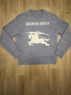 Grå stickad Burberry tröja med motiv - Säljer en grå stickad tröja från Burberry med ett stort vitt motiv av en riddare på häst och Burberry-loggan framtill. Tröjan har rund hals, ribbade muddar och är långärmad. Perfekt för dig som gillar klassiska designer med en twist.