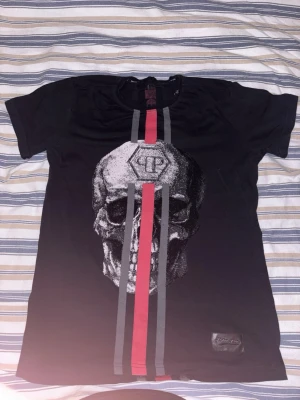 Svart t-shirt med döskalle från Philipp Plein - Cool svart t-shirt från Philipp Plein med stor döskalleprint och vertikala ränder i rött och grått på både fram- och baksida. Rund hals, korta ärmar och snyggt logopatch nertill. Materialet är mjuk bomull som känns skönt mot huden.