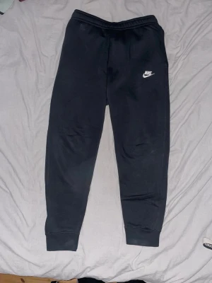Svarta mjukisbyxor från Nike, M - Svarta mjukisbyxor från Nike i storlek M med klassisk logga på vänster lår. Byxorna har elastisk midja och muddar vid bensluten, samt en bakficka. Perfekta för chill eller träning, tillverkade i mjukt bomullsmaterial. Väldigt bra skick 