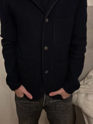 Mörkblå stickad kofta med knappar - Säljer en mörkblå stickad kofta med tre framfickor och knappar framtill. Koftan har lång ärm och klassisk krage, perfekt för lager-på-lager under kyliga dagar. Enkel och stilren design som funkar till jeans eller chinos.