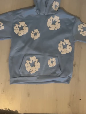 Denim Tears  - Blå hoodie med vitt blommigt mönster. Tillverkad i mjukt och bekvämt material med bra passform. Använd sparsamt och i mycket fint skick, inga hål eller fläckar. Perfekt för vardagsbruk.Fick som present har inte kommit till någon användning. Hör av dig vid frågor elller funderingar.