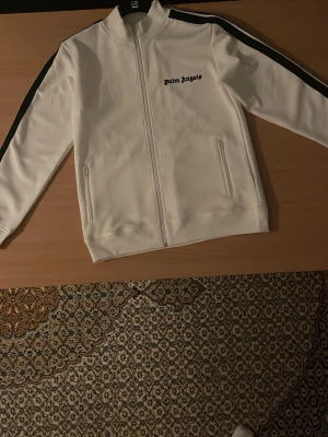 Vit Palm Angels track tröja - Säljer en fräsch vit track tröja från Palm Angels med svarta ränder längs ärmarna och logga på bröstet. Tröjan har hel dragkedja framtill, två fickor med dragkedja och en sportig, clean look. Perfekt för dig som gillar streetwear och vill sticka ut.