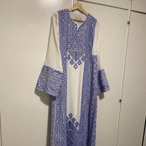 Vit långklänning med blått broderi - En palestinisk vit långklänning med detaljerat blått broderi och inslag av rött och gult. Klänningen har vida långärmar och ett geometriskt mönster längs hela plagget. Perfekt för dig som gillar traditionell design med modern twist. (Den har kommit till användning en kväll under 2 timmar). Köpte för några tusen och de drf lägsta priset kommer vara 1000kr.