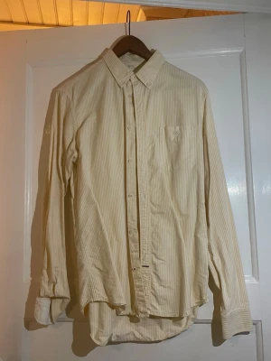 Randig skjorta från NN07 i bomull - Säljer en fräsch skjorta från NN07, modell Derek, i storlek M. Skjortan är vit med tunna ljusgula ränder, har klassisk krage, bröstficka och knappar framtill. Tillverkad i 100% bomull, perfekt för dig som gillar stilrena och softa plagg.
