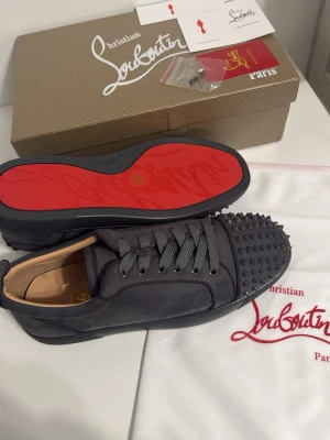 Gråa  Christian Louboutin skor - Säljer ett par gråa sneakers från Christian Louboutin med ikonisk röd sula och coola nitar på tån. Skorna har snörning och är tillverkade i skinn med en rund tå. Kommer med originalkartong, dustbag och äkthetskort.