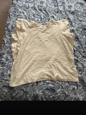 Beige blus med volang från Holly & Whyte/ Lindex  - Säljer en ljusbeige blus från Holly & Whyte by Lindex i storlek L. Blusen har korta ärmar med volangdetaljer och rund halsringning med knapp bak. Materialet är mjukt och känns som bomull, perfekt för vår och sommar.