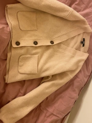 Beige kofta från Uniqlo, storlek S - Mysig beige kofta från Uniqlo i storlek S. Koftan har långa ärmar, två framfickor och stängs med fyra knappar. Ribbstickade detaljer vid muddar och nederkant ger en snygg touch. Perfekt för lager-på-lager under kyliga dagar.