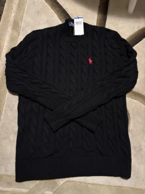 Svart kabelstickad tröja Polo Ralph Lauren - Kabelstickad svart tröja från Polo Ralph Lauren i bomull, med rund hals och ribbade muddar. Tröjan har ett rött broderat Polo-logo på bröstet och klassisk, chunky kabelstickning. Perfekt för dig som gillar stilrena och tidlösa plagg.