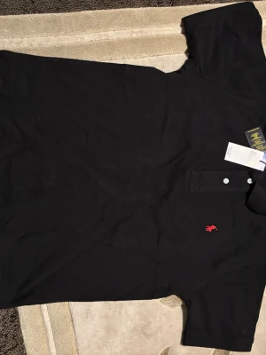 Svart piké från Polo Ralph Lauren - Säljer en svart pikétröja från Polo Ralph Lauren i klassisk passform. Tröjan har korta ärmar, krage och två vita knappar framtill. På bröstet finns det ikoniska röda Polo-broderiet. Materialet är bomull och känns mjukt och svalt mot huden.