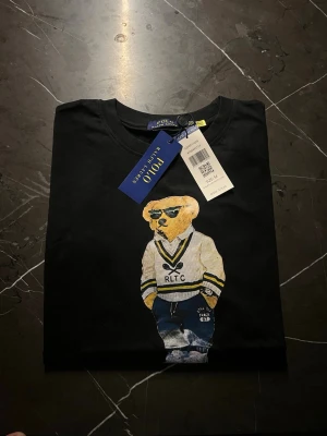 Svart Polo Bear t-shirt från Ralph Lauren - Svart t-shirt från Polo Ralph Lauren med ikoniskt Polo Bear-tryck framtill. Björnen har coola solglasögon och sportig outfit. Rund hals och korta ärmar. Materialet är mjuk bomull och trycket har vita, gula och blå detaljer. Perfekt för dig som gillar streetstyle och klassisk design.