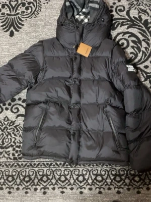 Svart dunjacka från Burberry - Säljer en svart dunjacka från Burberry med huva och klassiskt rutigt foder. Jackan har dragkedja och knappar framtill, två snedställda fickor med dragkedja och en patch med Burberry London England på ärmen. Perfekt för kalla dagar och riktigt snygg streetstil.