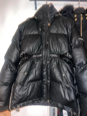 Manière De Voir jacka - Säljer en svart pufferjacka från Manière De Voir med glansiga detaljer och stora dragkedjor på sidorna. Jackan har hög krage, tryckknappar och fickor framtill. Perfekt för kalla dagar.