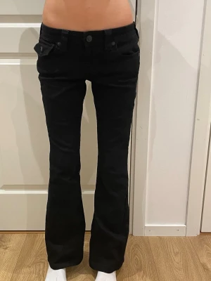 Svarta bootcut jeans från True Religion - Snygga svarta jeans från True Religion med bootcut-snitt och låg midja. Byxorna har klassiska fickor fram och bak med lock och knappdetaljer, samt en tydlig logopatch bak. Materialet är jeans och passformen är normal.Storleken är 26,skickar inom 1-2 dagar!💓