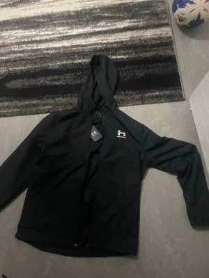 Svart vindjacka från Under Armour - Svart vindjacka från Under Armour med huva och dragkedja framtill. Den är oanvänd och har lappen på. Anledningen till att jag säljer den är för att jag tappade kvittot. Den kostade 1229kr, säljer den för 650kr, en mycket bra deal för dig. 