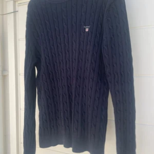 Mörkblå kabelstickad tröja från GANT - Säljer en mörkblå kabelstickad tröja från GANT i storlek L. Tröjan har rund hals, lång ärm och ett broderat GANT-logo på bröstet. Perfekt för dig som gillar klassisk stil med snygga detaljer. Materialet är mjukt och stickat för extra komfort.