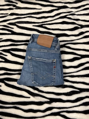 Replay Grover jeans  - Säljer ett par Replay Grover jeans i klassisk blå tvätt med knappar i gylfen. Jeansen har fem fickor, snygga detaljer och en skinnpatch bak i midjan. Materialet är robust! En defekt vid grenen ett hål kan nog sy ihop men ingen passion här pga försmå hör av dig vid minsta fundering 😌