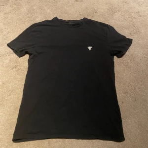 Svart t-shirt från Guess, super slim - Svart t-shirt från Guess med klassisk rund hals och diskret logga på bröstet. Modellen är super slim och sitter tajt mot kroppen. Tillverkad i mjuk bomull som känns skön hela dagen. Perfekt för dig som gillar stilrena och enkla plagg med en touch av street.