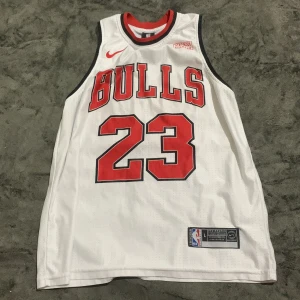 Chicago Bulls Jordan 23 baskettröja - Snygg Chicago Bulls baskettröja från Nike med nummer 23 och Jordan på ryggen. Tröjan är vit med röda och svarta detaljer, har ärmlös design och meshmaterial som andas. Perfekt för dig som vill ha en ikonisk sportlook.