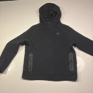 Svart Nike hoodie med dragkedja - Svart hoodie från Nike med hel dragkedja framtill och två praktiska fickor med dragkedja. Tröjan har huva och är tillverkad i mjuk bomullsblandning. Perfekt för en sportig och avslappnad stil.