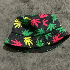 Svart bucket hat med färgglada blad - Cool svart bucket hat med färgglada blad i grönt, gult och rött. Hatten har ett avslappnat snitt och är tillverkad i bomull. Perfekt accessoar för att sticka ut och ge din outfit en unik vibe.