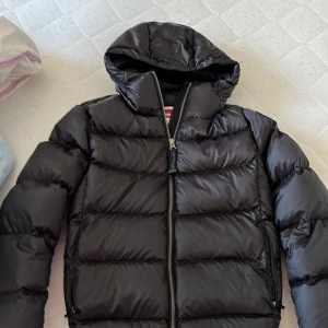 Svart pufferjacka från Parajumpers - Säljer en svart pufferjacka från Parajumpers med huva och dragkedja framtill. Jackan har klassisk quiltad design och logotyp på ärmen. Perfekt för kalla dagar och riktigt snygg streetstil.