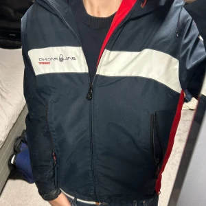 Sail Racing vindjacka - Snygg marinblå vindjacka från Sail Racing med röda och vita detaljer. Jackan har huva, dragkedja framtill, två fickor med dragkedja och logga på bröstet och ryggen. Perfekt för dig som gillar sportig stil och vill sticka ut med coola färgblock. Priset kan diskuteras