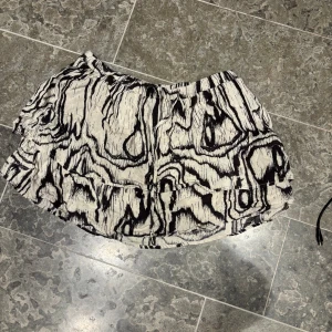 Svartvit kortkjol med abstrakt mönster - Kort kjol i svart och vitt med ett unikt abstrakt mönster som påminner om träådring. Kjolen har volangdetalj och resår i midjan för en bekväm passform. Perfekt för dig som vill sticka ut med en cool och annorlunda stil.